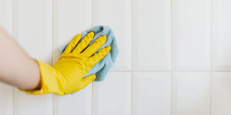 A person in yellow gloves actively cleans a tile wall, illustrating a hard water stain removal service in action.