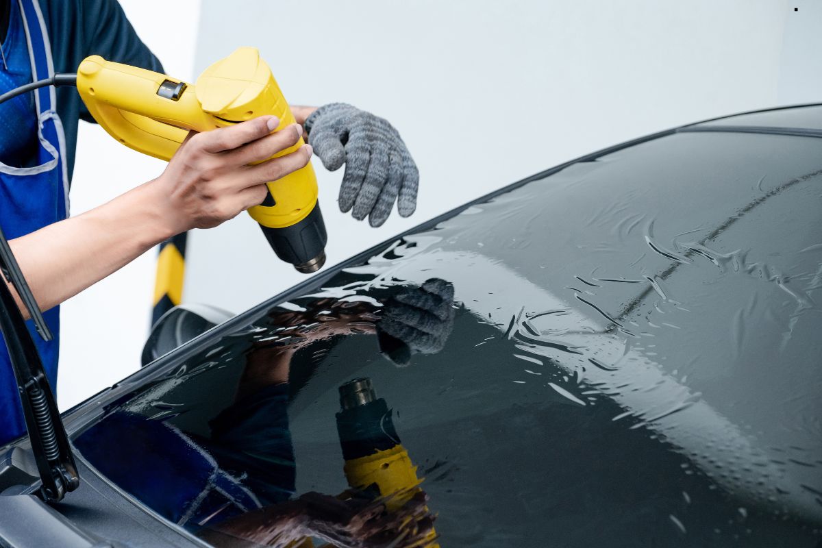 A technician uses a yellow heat gun on a car window, smoothing dark window tint with gloved hands, reflecting on the glass surface.