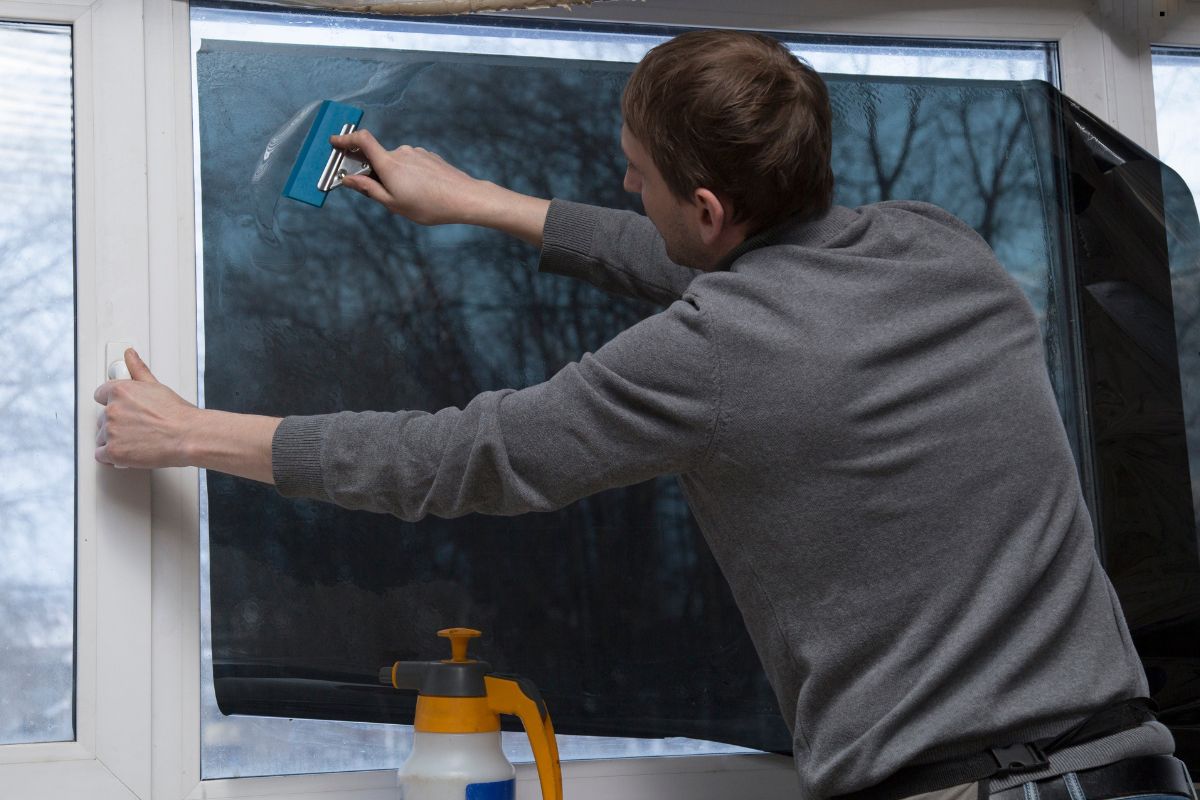 A person is applying window film to a pane, using a blue scraper to smooth out air bubbles, while a spray bottle sits nearby.