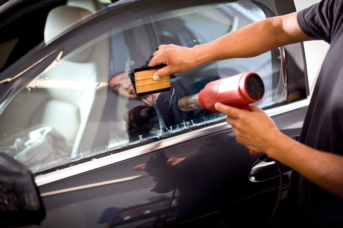 A technician applies film to a car window using a squeegee and a heat gun, ensuring a smooth finish.